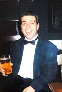 Smiling person in a tuxedo holding a drink.