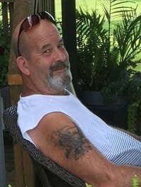 Smiling man with sunglasses and tattoo sitting outside.