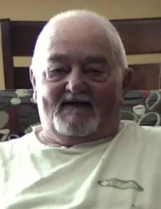 Elderly man smiling, sitting indoors on a chair.