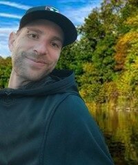 Person smiling outdoors near a river and trees.