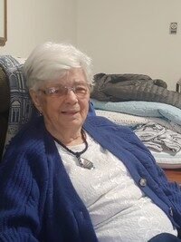 Elderly woman sitting indoors, wearing glasses and sweater.