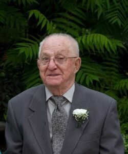 Elderly man in suit with boutonniere, greenery background.