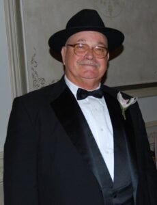 Man in tuxedo and hat smiling indoors.