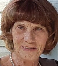 Elderly woman smiling outdoors with short brown hair.