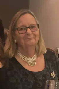 Woman wearing glasses and pearls, seated indoors.