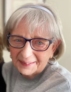 Elderly woman wearing glasses and gray sweater smiling.
