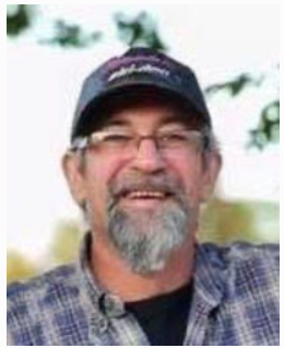 Smiling man wearing cap and glasses outdoors.