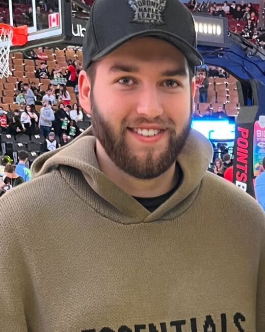 Man in arena wearing hoodie and cap, smiling.