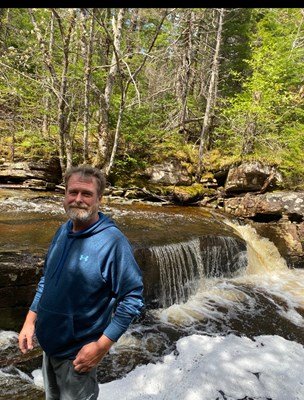 Man stands by forest waterfall in blue hoodie.