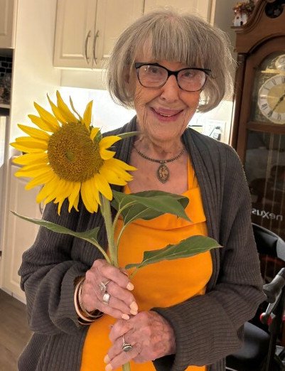 Elderly woman holding a sunflower in kitchen.