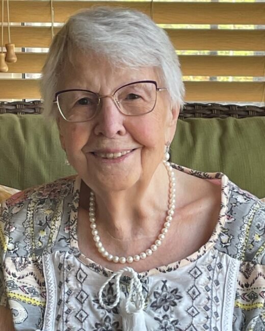 Smiling elderly woman with glasses and pearl necklace.