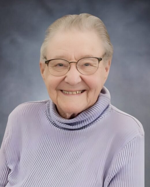 Smiling elderly person wearing glasses and purple sweater.