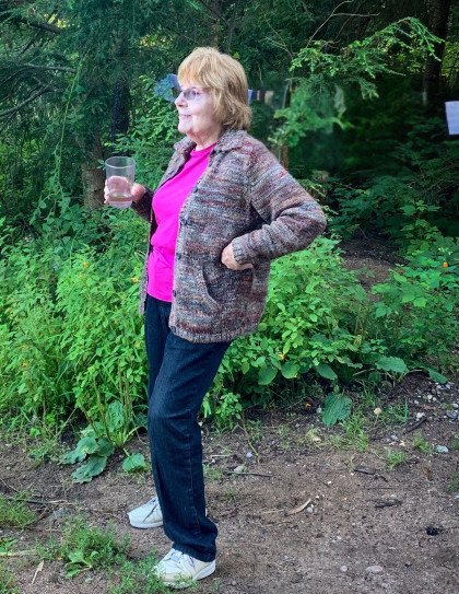 Woman in garden holding glass, wearing sweater and jeans.