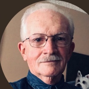 Elderly man with glasses and moustache smiling indoors.