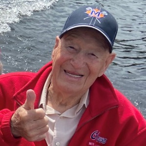 Smiling person in red jacket, thumbs up by water.