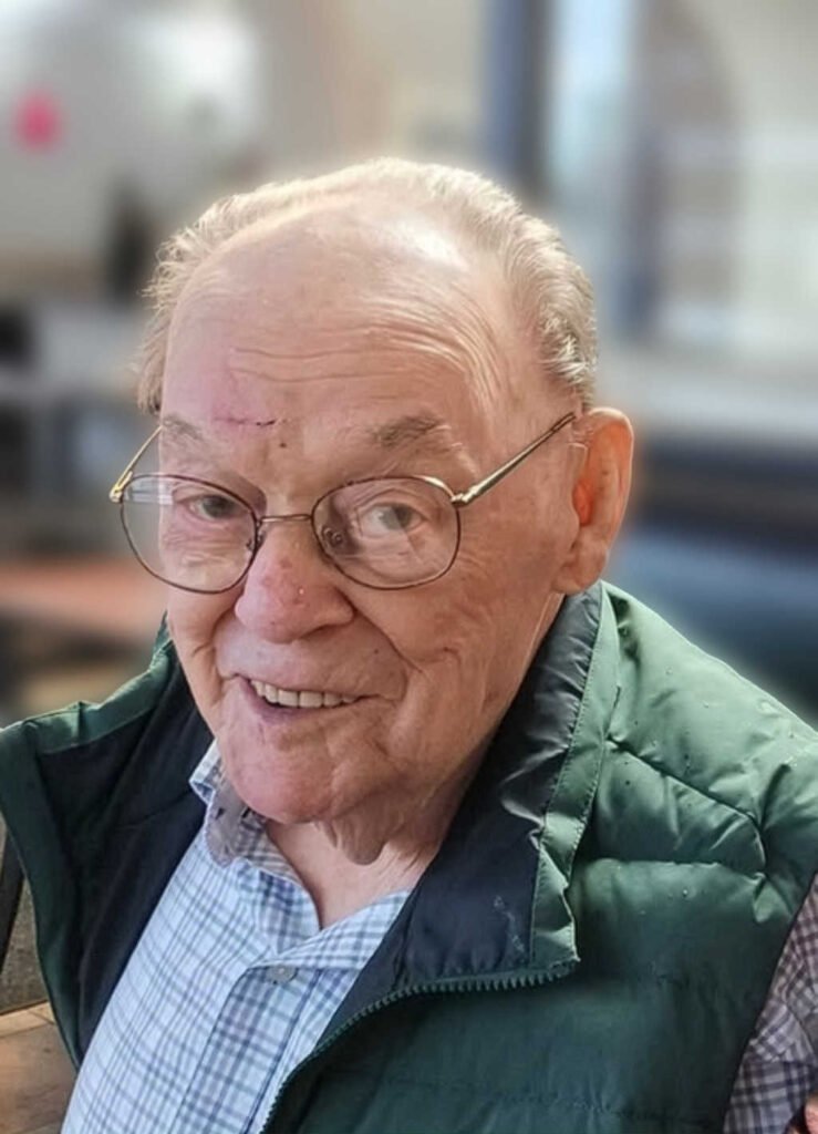Elderly man smiling with glasses and green jacket.