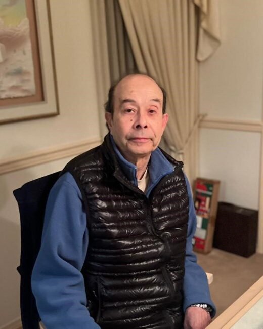 Man sitting indoors wearing blue and black jacket.