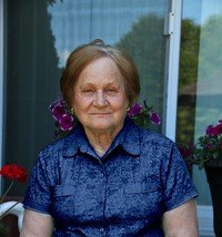 Elderly woman sitting outdoors with flowers nearby.