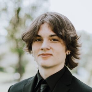 Person smiling in formal black suit outdoors.