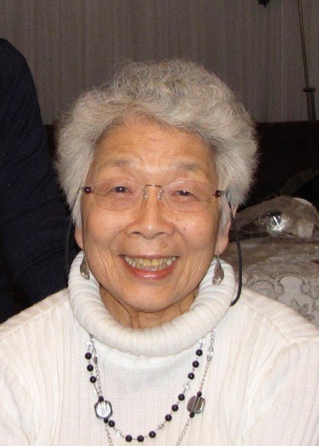 Elderly woman smiling indoors wearing glasses and necklace.