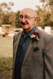 Elderly man in suit with boutonniere, smiling outside.