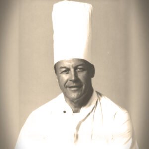 Chef wearing tall white hat and uniform.
