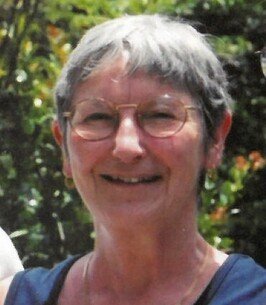 Elderly woman smiling outdoors with greenery behind.