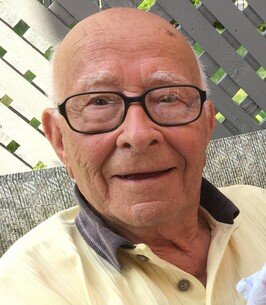 Elderly man smiling outdoors wearing glasses.