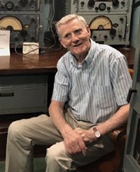 Elderly man in radio equipment room smiling seated.