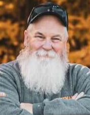 Smiling man with white beard and sunglasses outdoors.