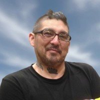 Smiling man with glasses and tattoos under blue sky.