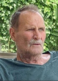 Older man sitting outdoors with greenery background.