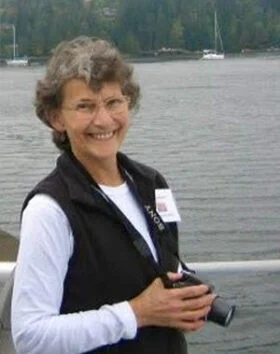 Person smiling near water, holding a camera.