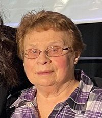 Elderly woman wearing glasses and plaid shirt, smiling.