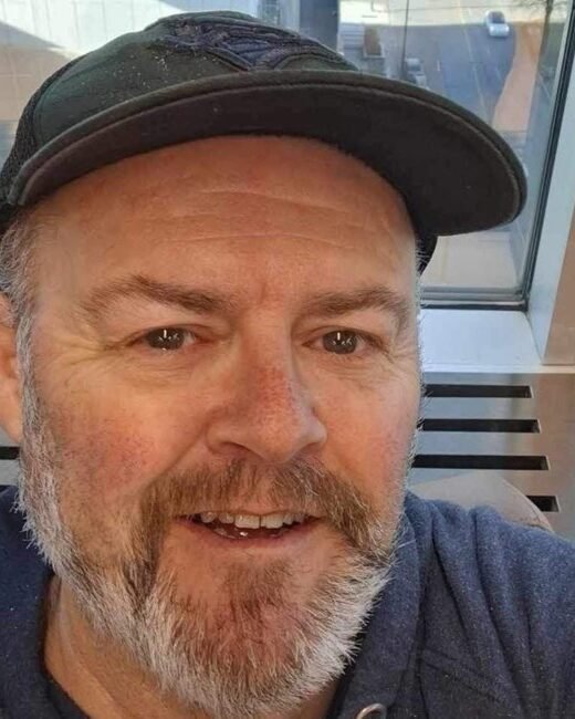 Man wearing a hat smiling indoors near window.