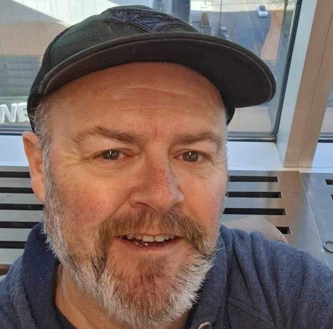 Man wearing a hat smiling indoors near window.
