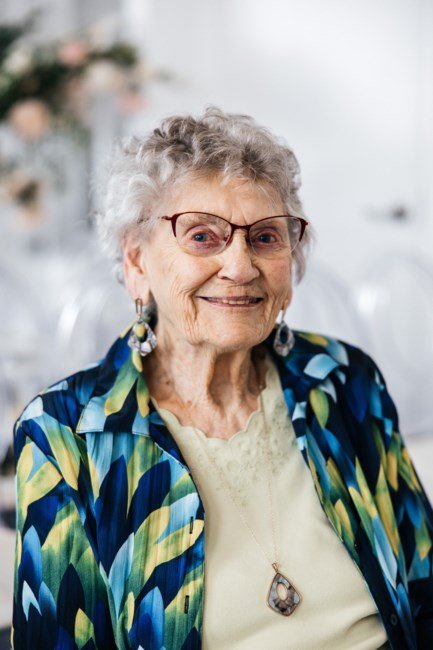 Elderly woman smiling in colorful blouse and glasses.