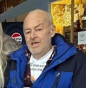 Older man indoors wearing blue jacket and plaid shirt.