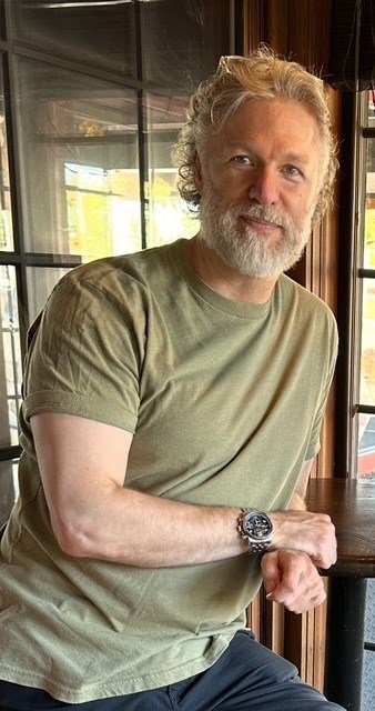 Man with beard sitting indoors with window background.