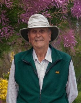 Elderly man outdoors wearing hat and green vest.