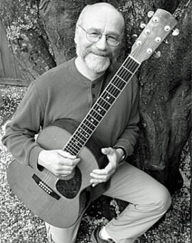 Man holding an acoustic guitar, smiling outdoors.