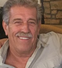 Smiling man sitting indoors with stone wall backdrop.