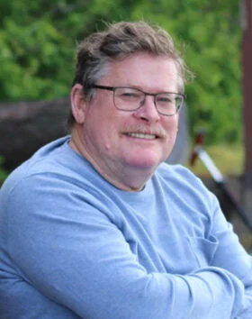 Smiling man sitting outdoors in blue sweater.
