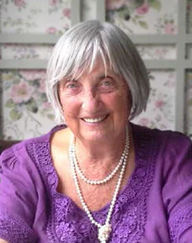 Smiling elderly woman in purple blouse with pearls.