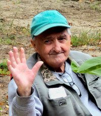 Smiling elderly man waving outdoors with green cap.