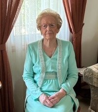 Elderly woman seated by curtain in a teal dress.