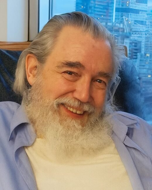 Smiling man with beard, sitting indoors by window.