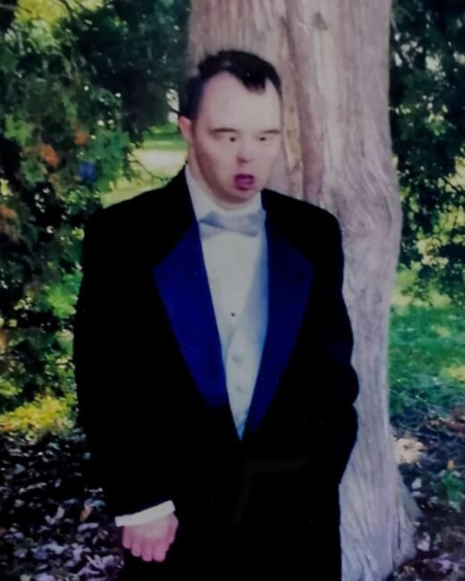 Person in formal suit standing by trees outdoors.