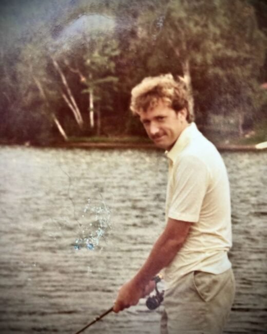Man fishing by a calm lakeside with trees.