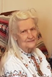 Elderly woman in embroidered blouse sitting indoors.